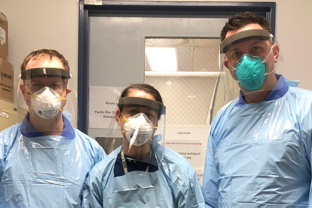 Three people in personal protective equipment stand in a lab doorway.