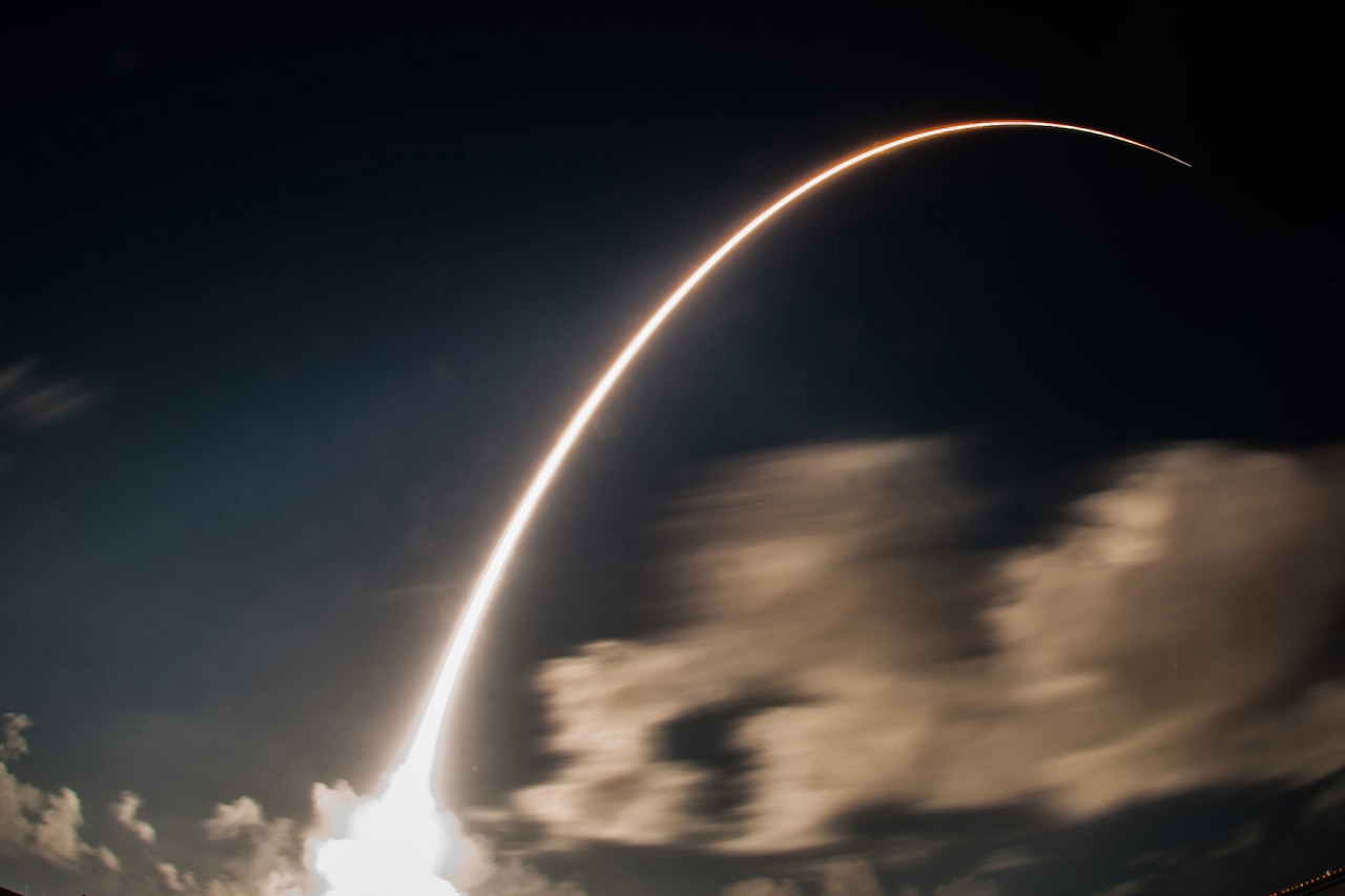 A rocket arcs into the night sky leaving behind a contrail of light.