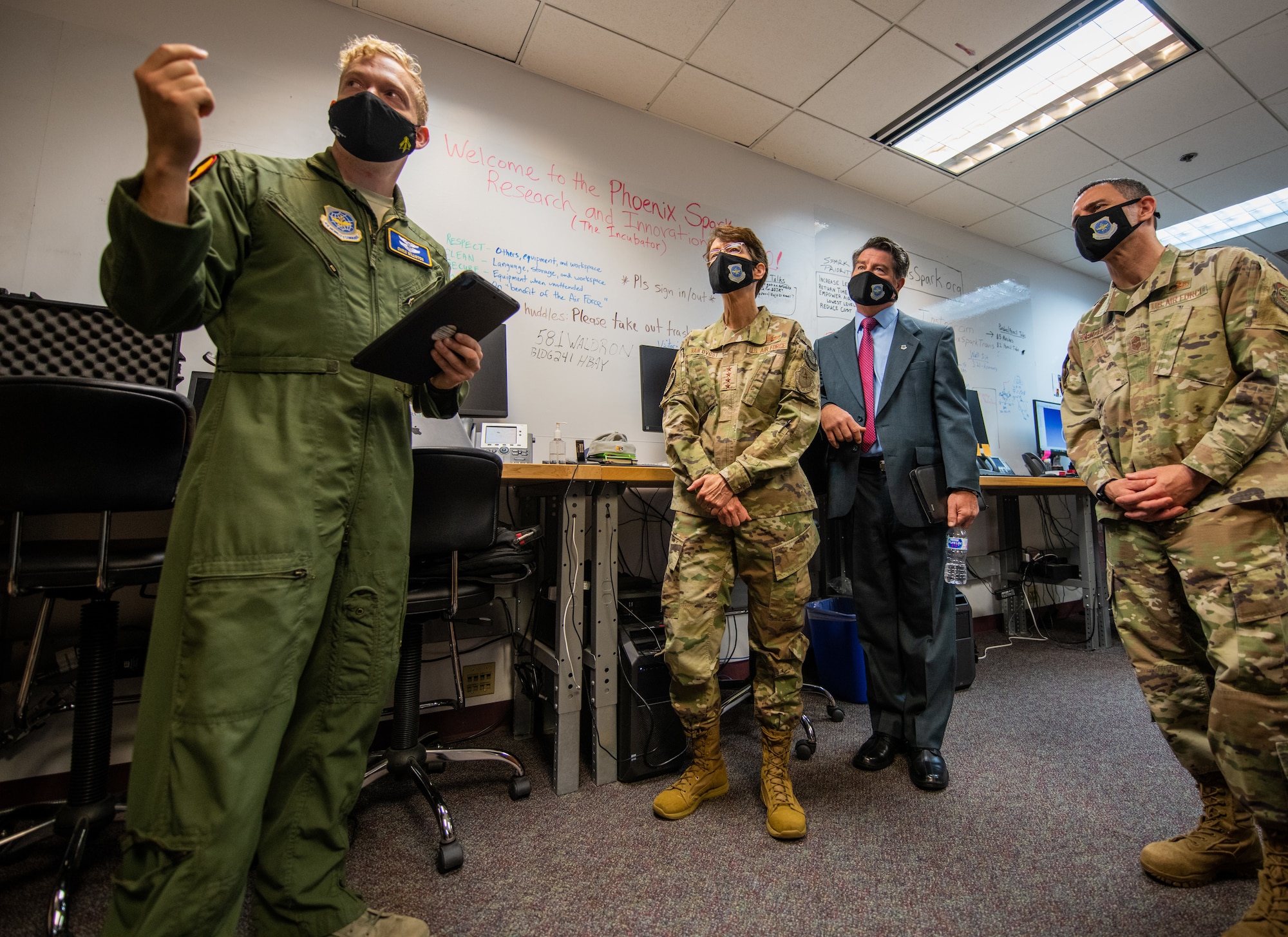 Military members and one man in a suit stand inside a room with fluorescent lights and a carpeted floor. They are all wearing masks.