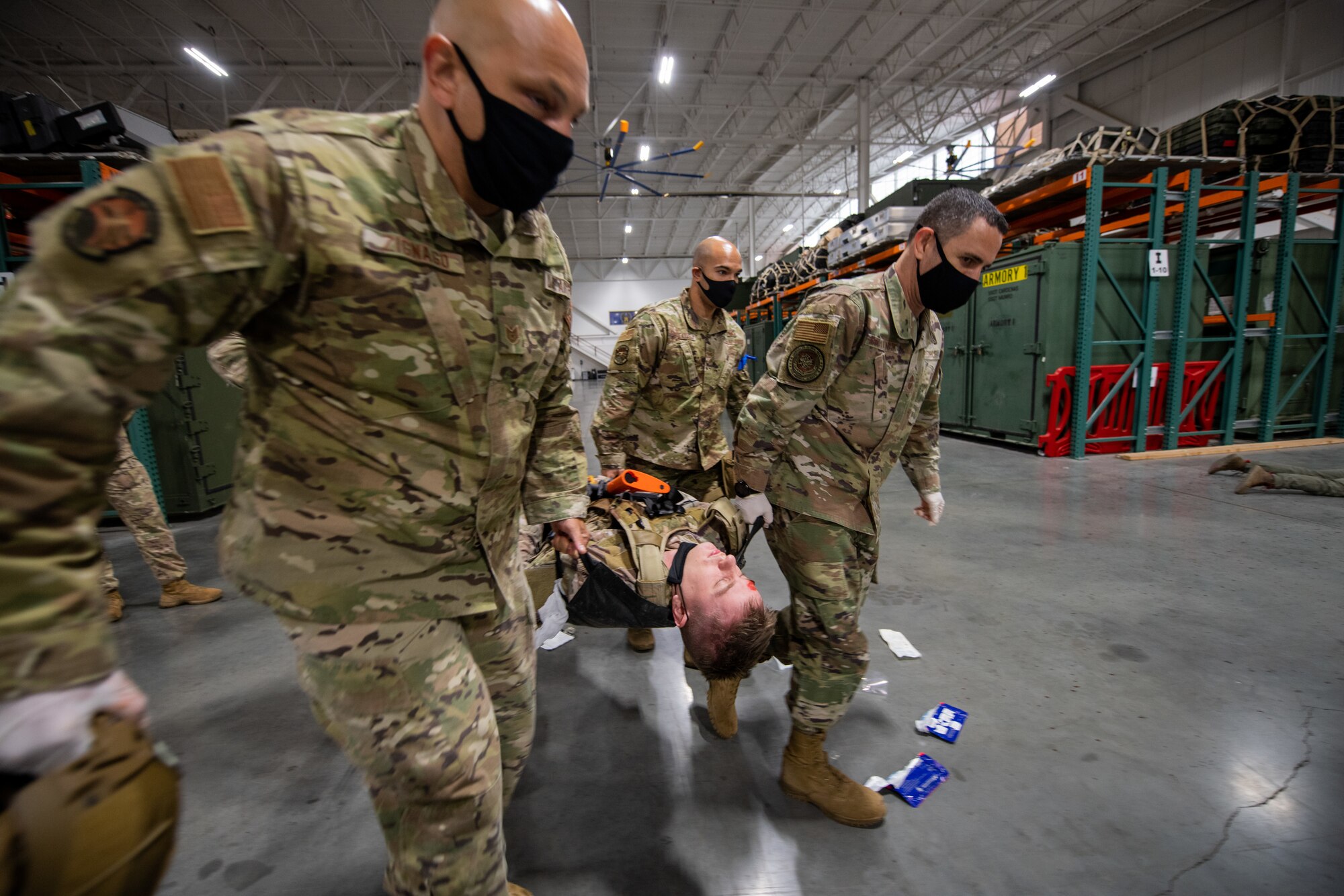 Three military members help to hand-carry a fourth on a stretcher inside of a warehouse with fluorescent lights