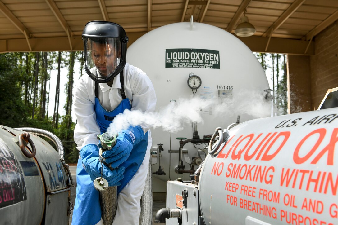 An airman in protective gear holds a hose that has smoke coming out of it beside a tank of liquid oxygen.