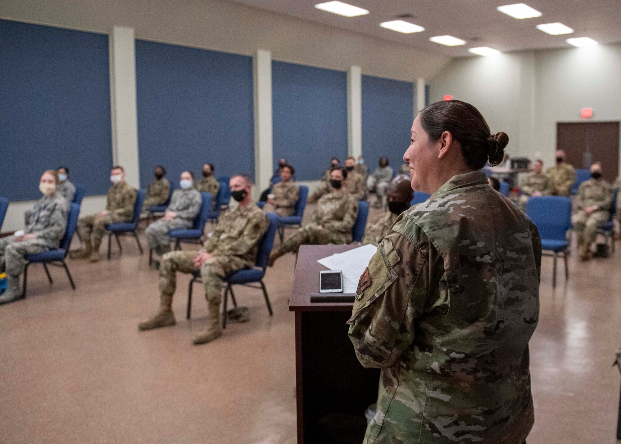 A female Airman stands behind a podium, smiling, in front of an audience