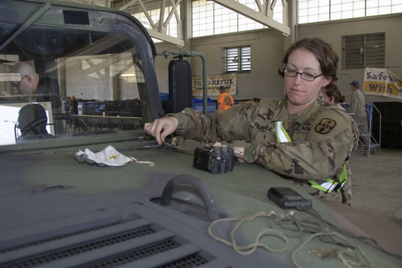 A female service member looks at some equipment that sits on the hood of a military vehicle.