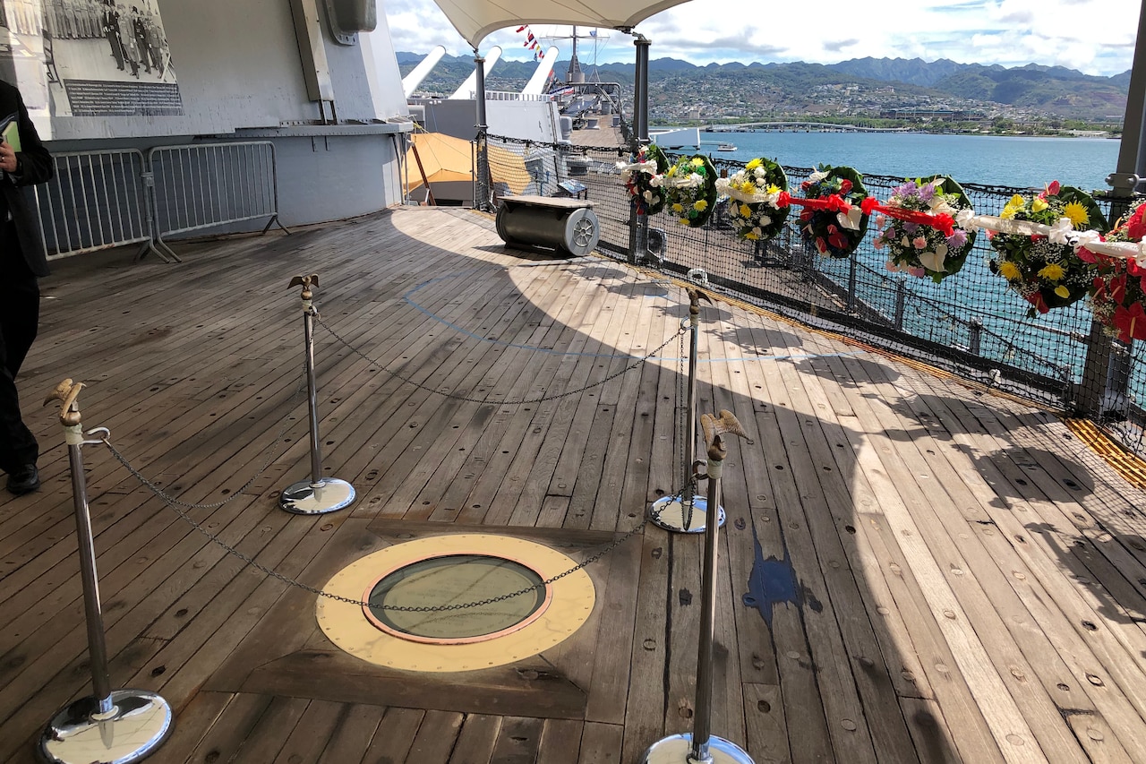 A plaque on the deck of a battleship surrounded by floral tributes.