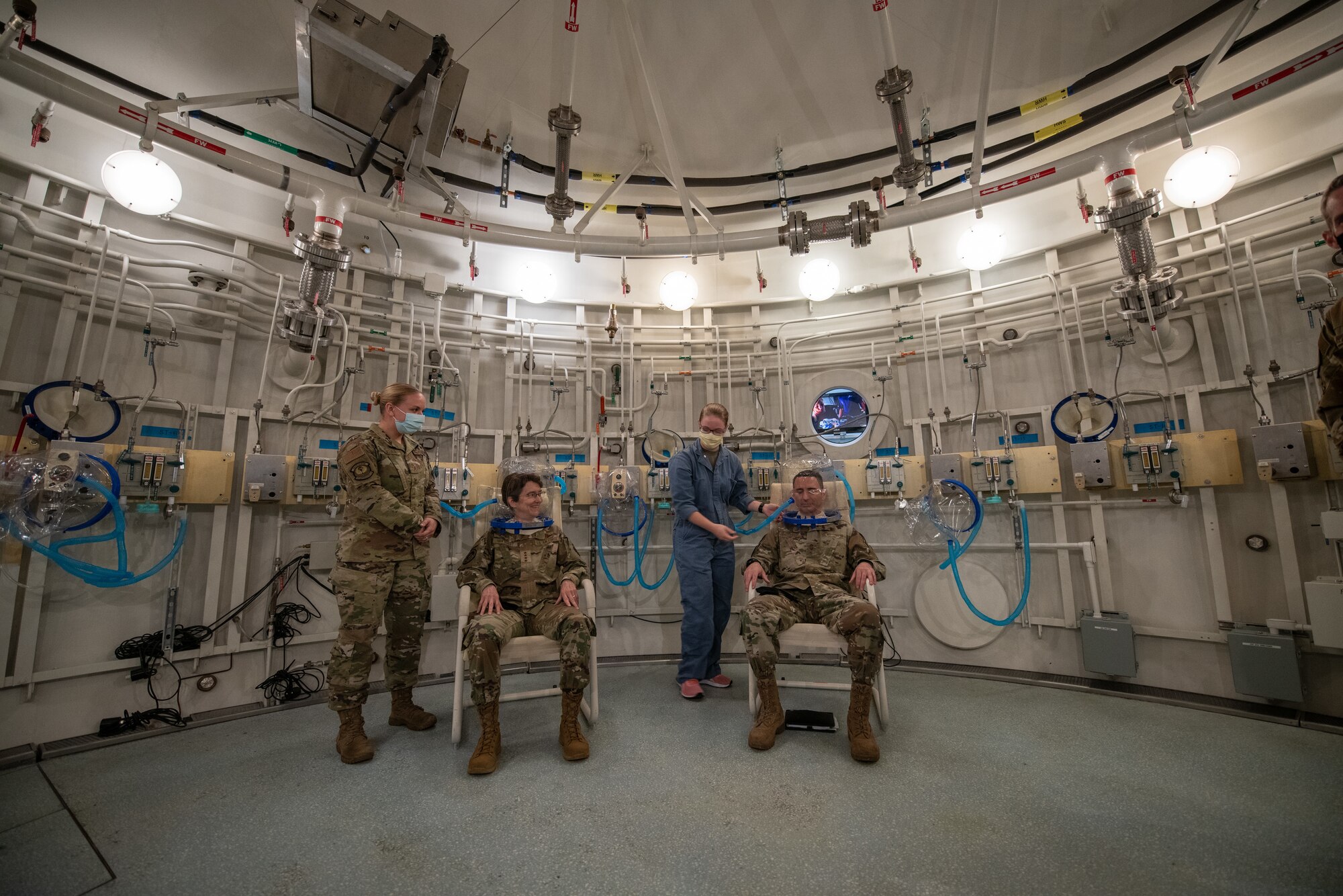 Three military members sit inside a large, circular room with a medic. All are wearing face masks. There are pipes and valves on the white walls.