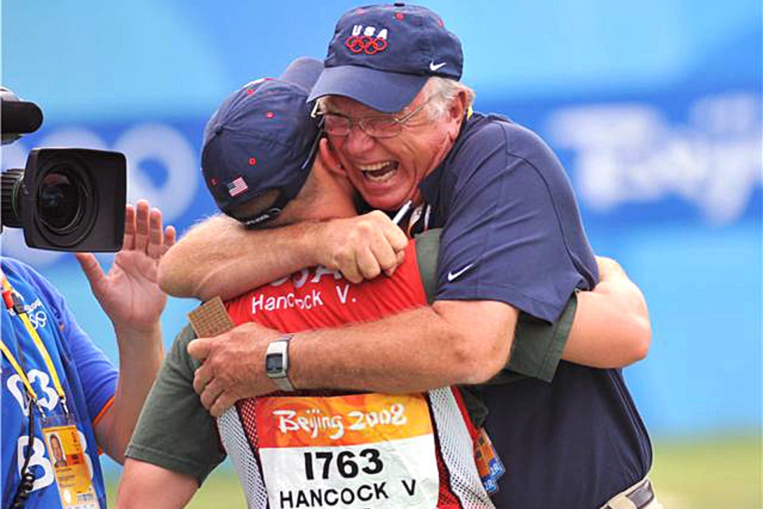 An older man hugs a man wearing a vest with the words Beijing 2008 Hancock V. USA.