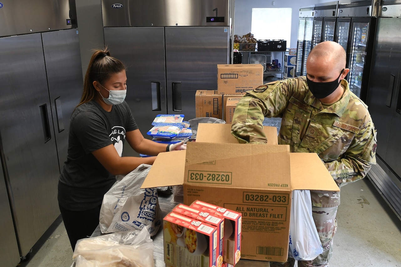 A service member and a woman wearing a food bank tee shirt pack a box with food.