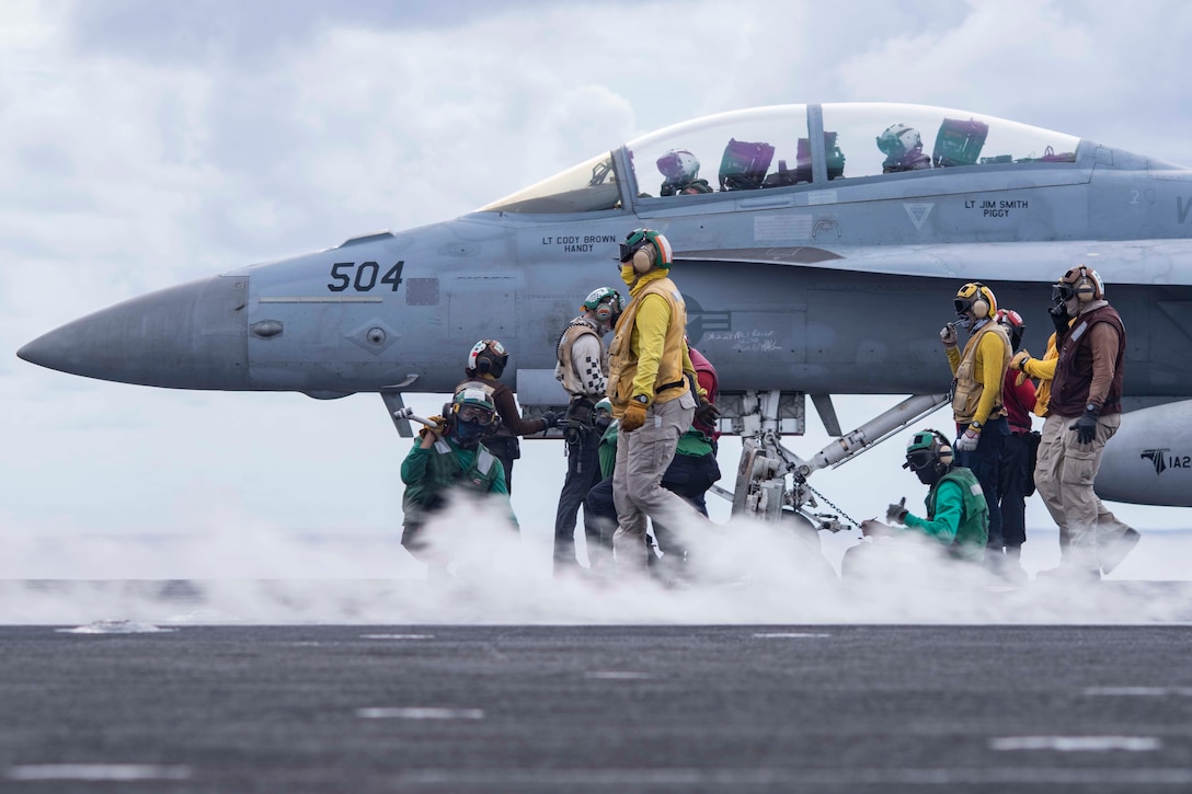 Sailors work on an aircraft as smoke passes by.