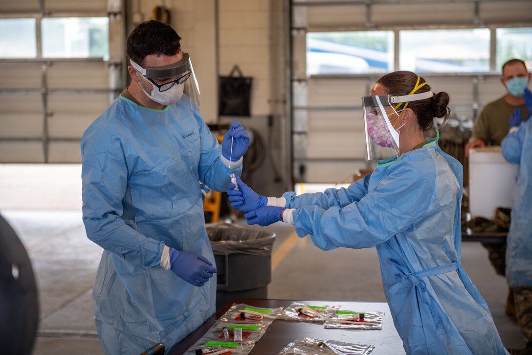 A man dressed in protective gear prepares to put a swab into a vial held by a woman, who is also dressed in protective gear.