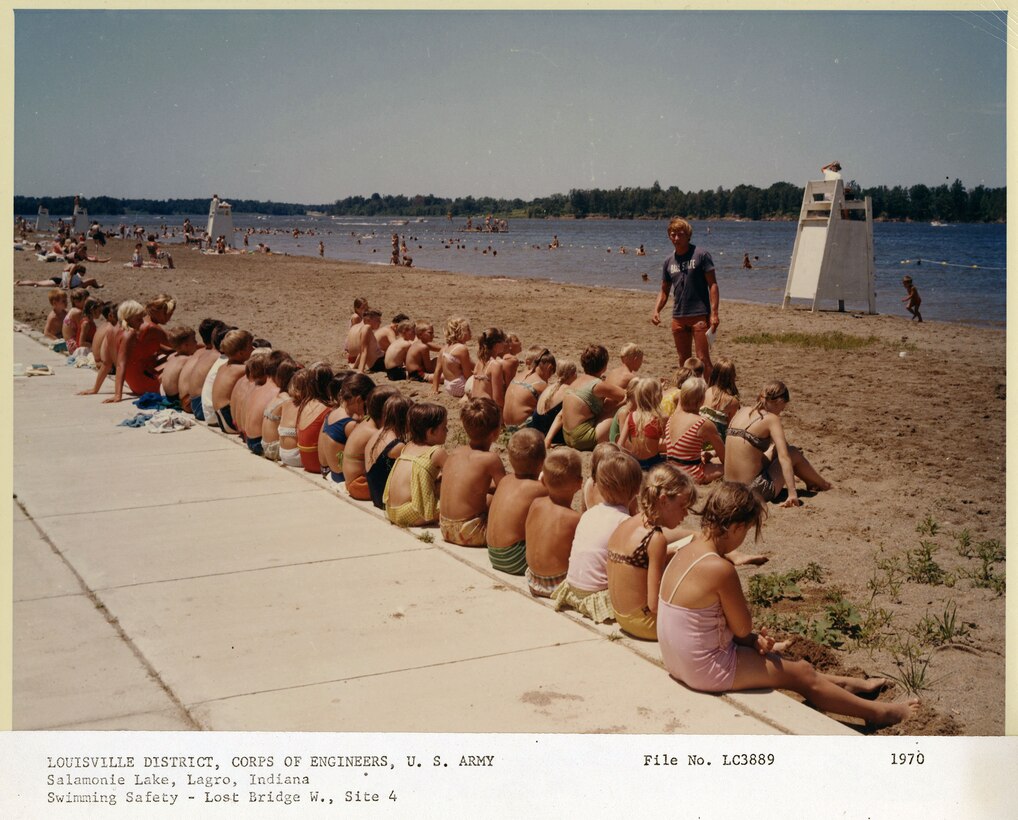 Children sit in rows on the sand listening to a male lifeguard give a swimming safety lesson with lake in the background