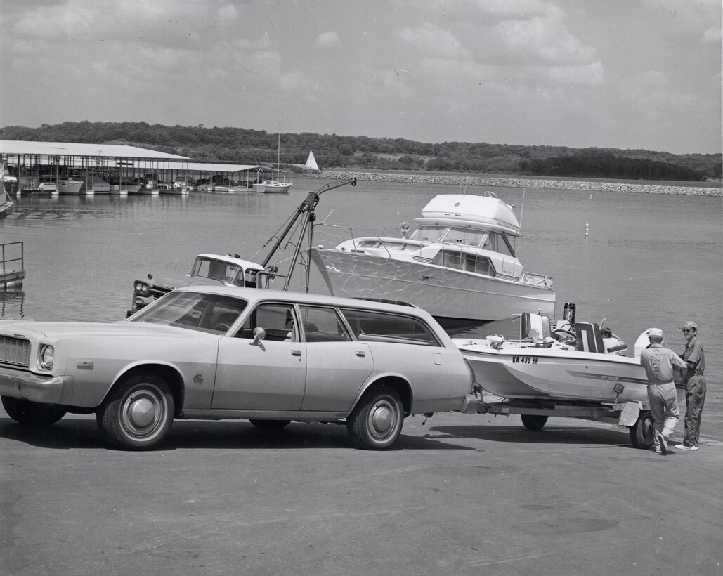A station wagon backs a small boat down a ramp to the shoreline as two men watch