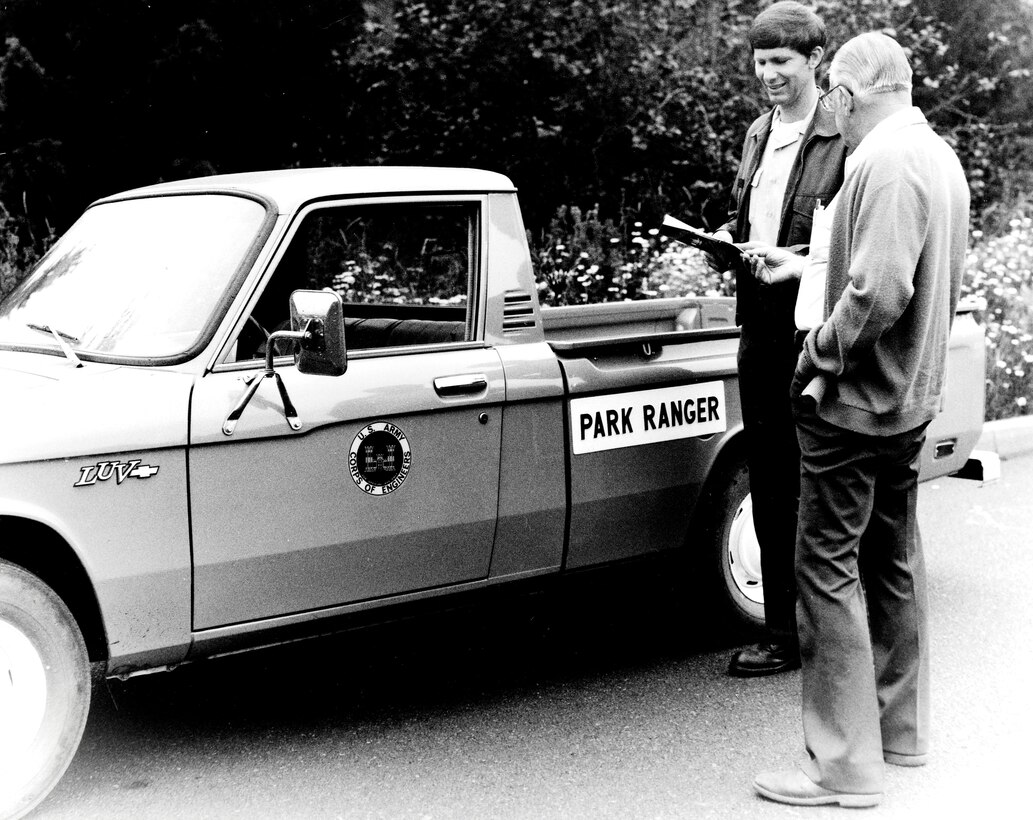 A park ranger and a visitor stand outside a small pickup truck with the Corps logo and park ranger decals