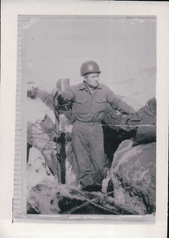 A helmet-clad soldier stands by a bunker with his hand on a metal pole.