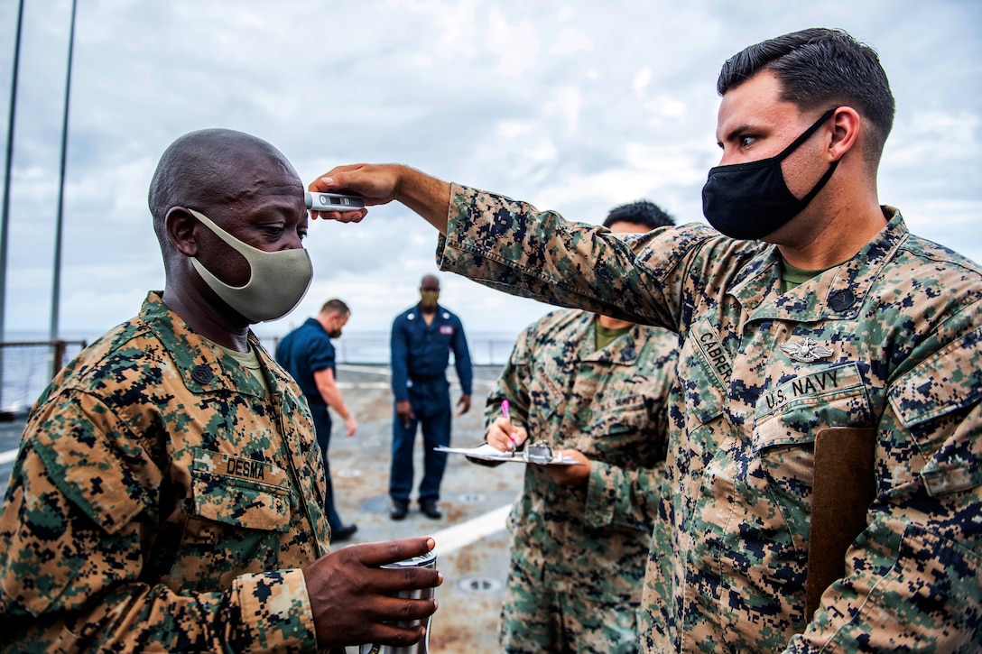A sailor wearing a face mask takes the temperature of another service member wearing a face mask.