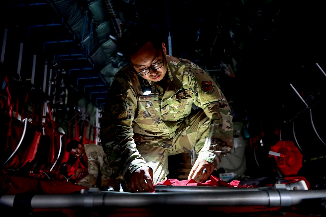 An airman kneels  with a lit flashlight cradled between his shoulder and face while doing maintenance work inside a dark aircraft.