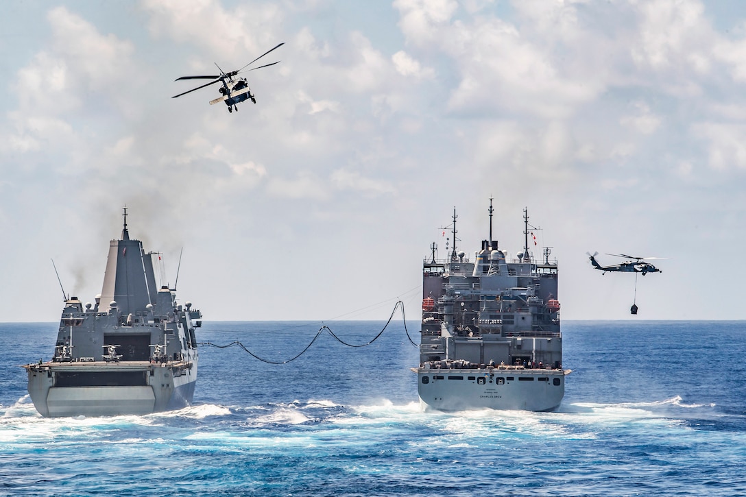Two ships sail next to each other as helicopters fly above.