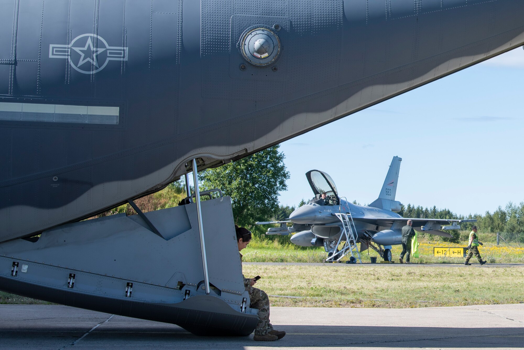 An Airman with the 352d Special Operations Aircraft Maintenance Squadron sits on the back ramp of an MC-130J Commando II, based out of RAF Mildenhall, U.K., as a Royal Norwegian Air Force pilot prepares to depart his F-16 Fighting Falcon during a training mission at Rygge Air Station, Norway, August 25, 2020. Integration with the Norwegian Air Force allowed the 352 SOW to enhance and strengthen bonds with our partner nation and further secure the strategic high-north region. The exercise provided training for 352d Special Operations Wing members on capabilities such as personnel recov-ery, forward area refueling point, aerial refueling, maritime craft delivery system, and fast rope training. (U.S. Air Force photo by Staff Sgt. Michael Washburn)