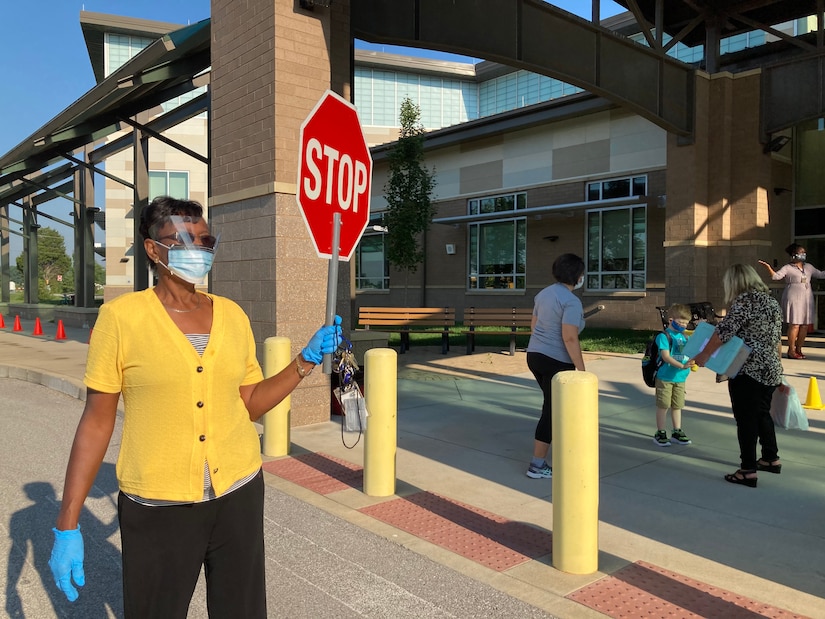 A teacher wearing a face mask and shield holds a stop sign.