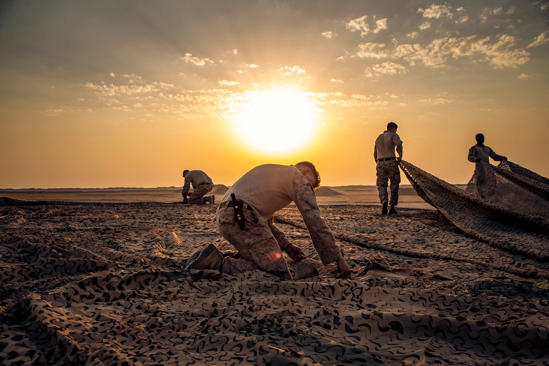 Marines work on a large canopy laid out flat on the ground on desert terrain, with a low sun and orange sky in the background.