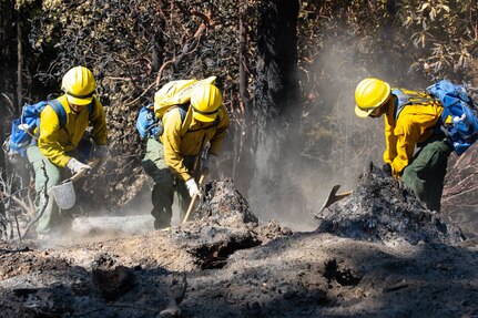 Three soldiers in firefighting gear dig in the dirt.