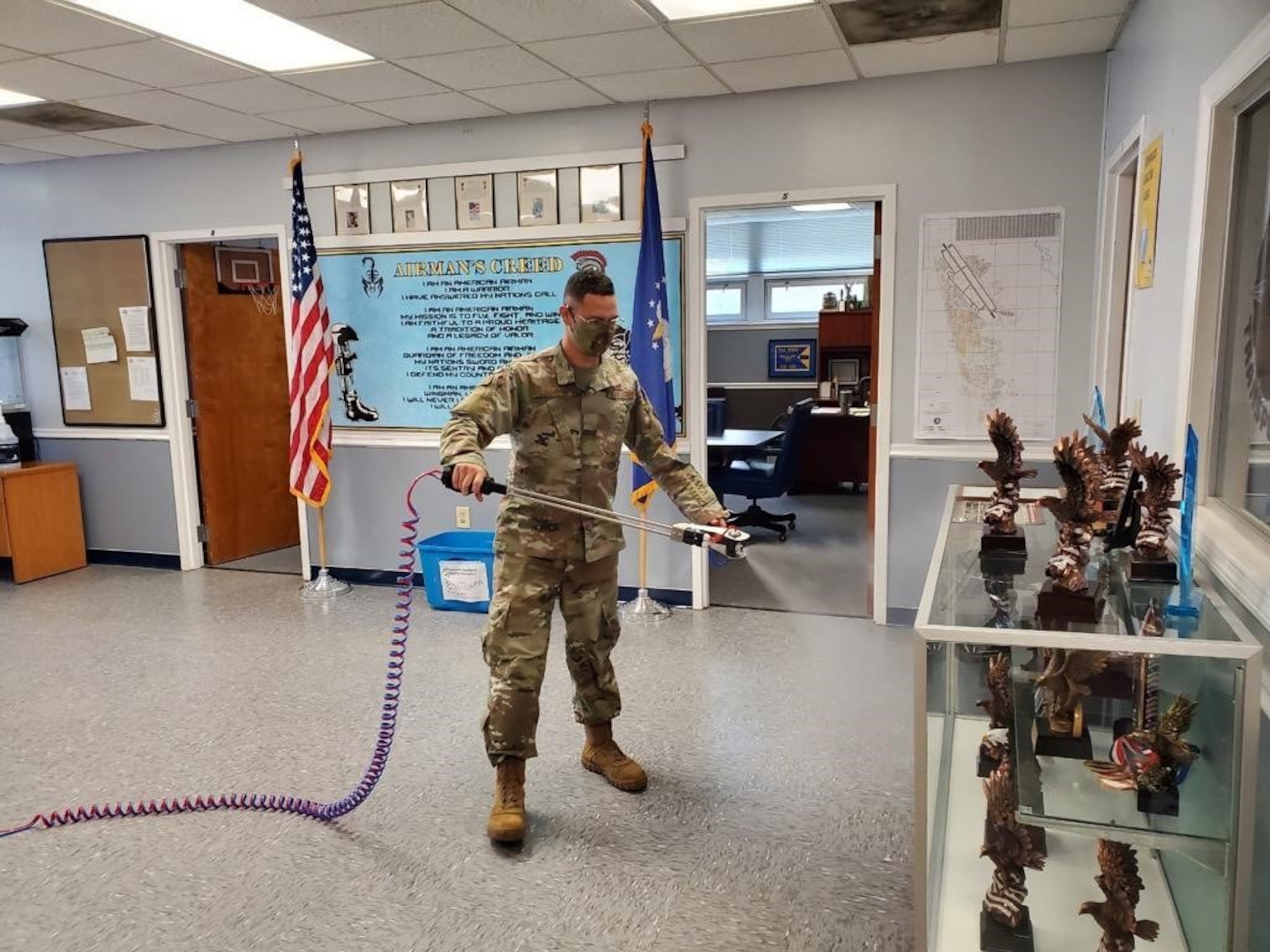 Photo shows an Airman indoors spraying disinfectant around his work area.