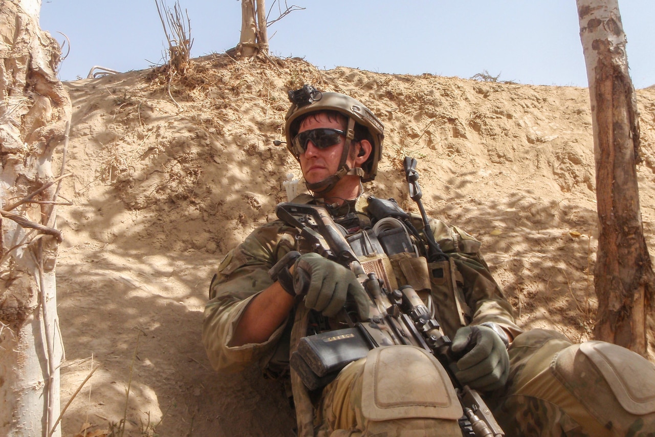 A soldier holding a weapon sits in a desert like area.