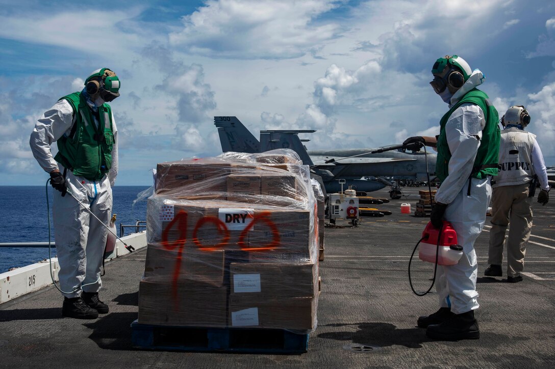 Two airmen spray disinfectant on a stack of boxes.
