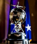 A trophy with a silver globe and lightning bolts displayed on top of 2 black tier circles with military flags draped in the back ground.
