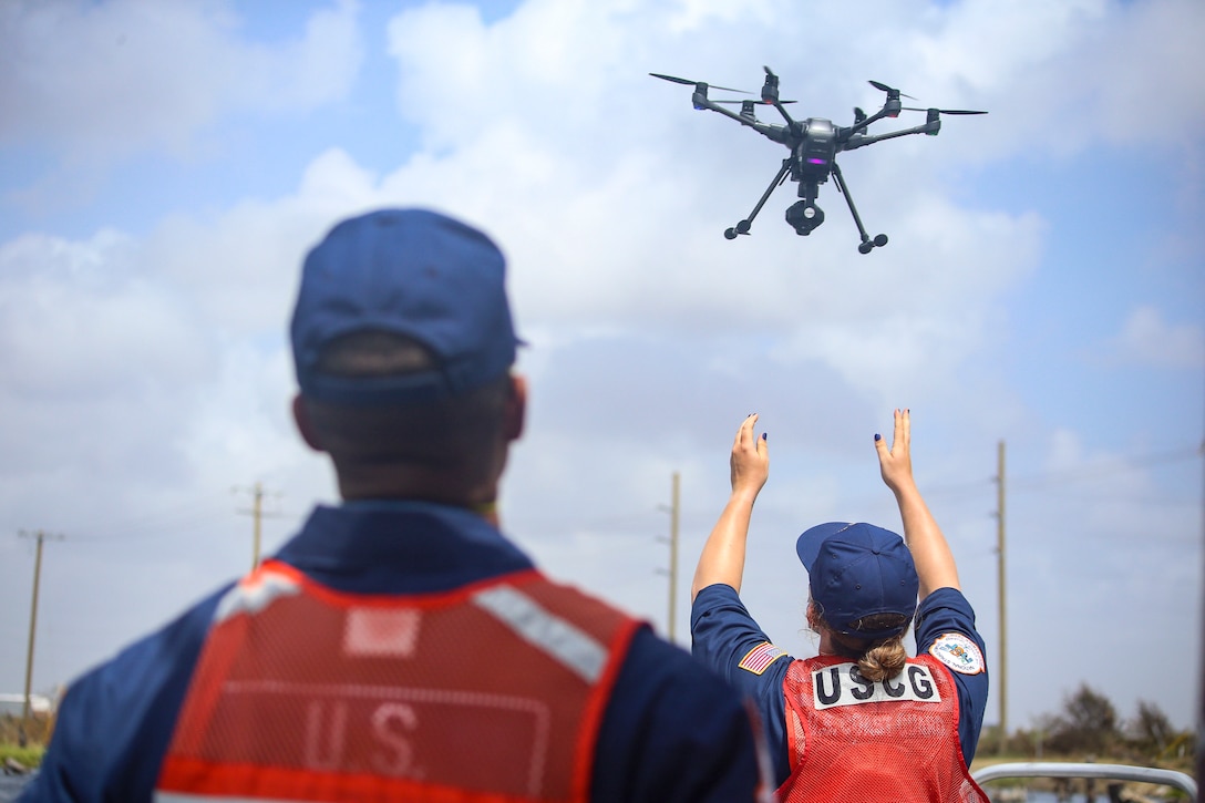 Two Coast Guardsman, shown from behind, watch as drone hovers in the air. One has her arms up as though she launched it.