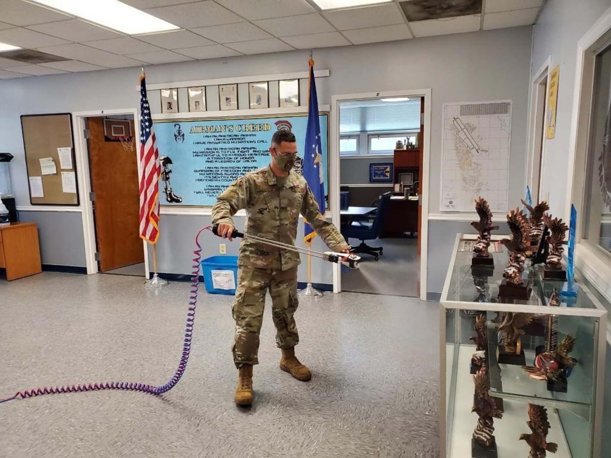 Photo shows an Airman indoors spraying disinfectant around his work area.