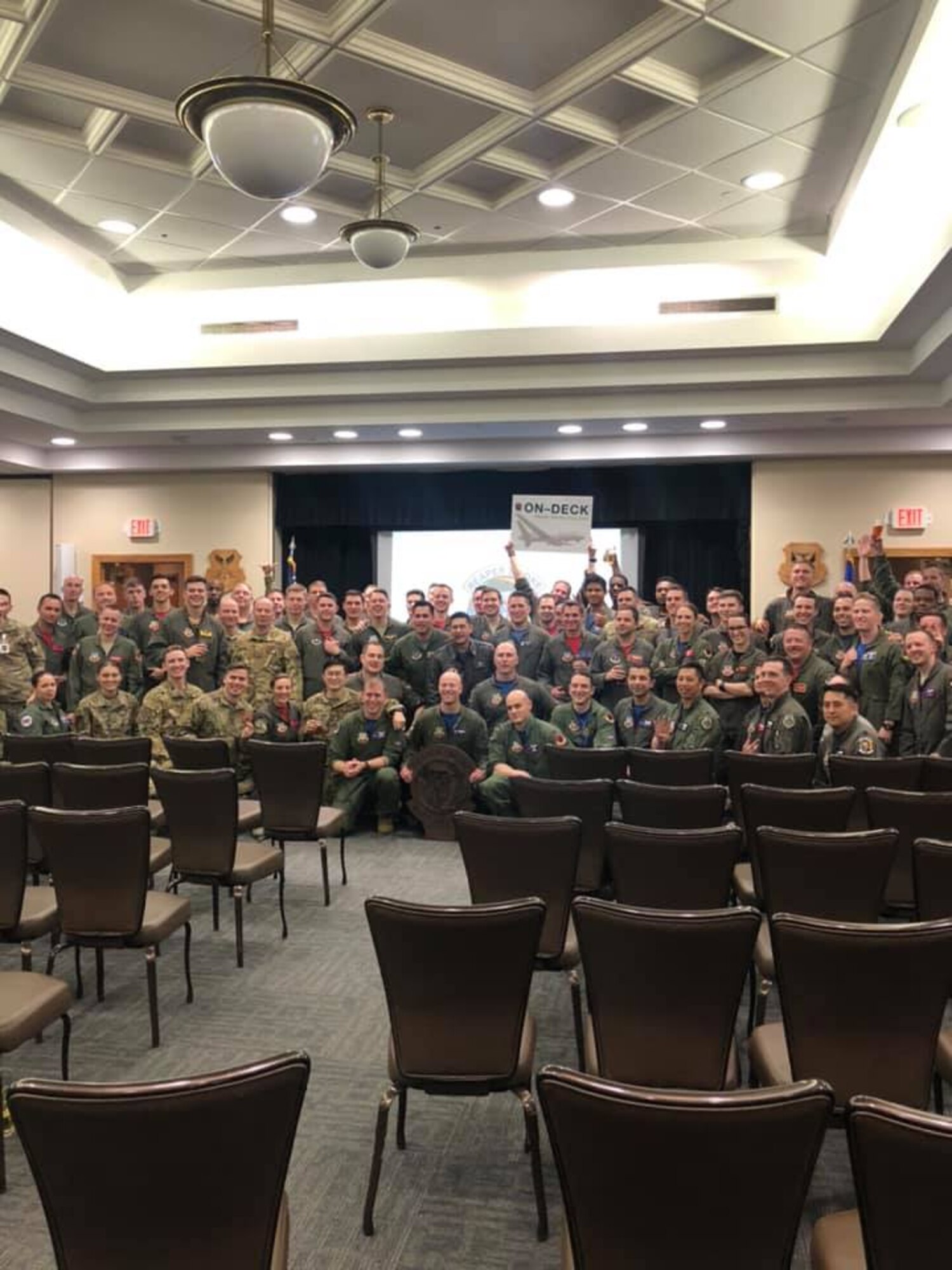 68 military members, in uniform, pose indoors at a lecture hall and holding a squadron emblem in the center of the photo with all smiles in celebration.