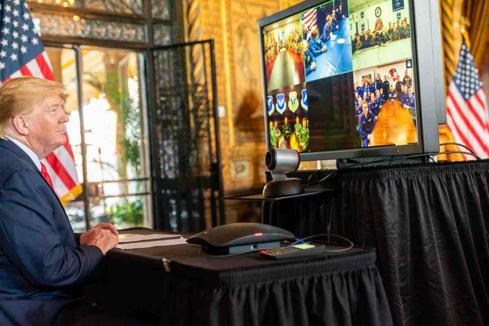 President Trump sitting at a table and looking at a monitor with images from 5 different locations of military members ranging from 5 to 15 people looking back at the president while having a conversation.