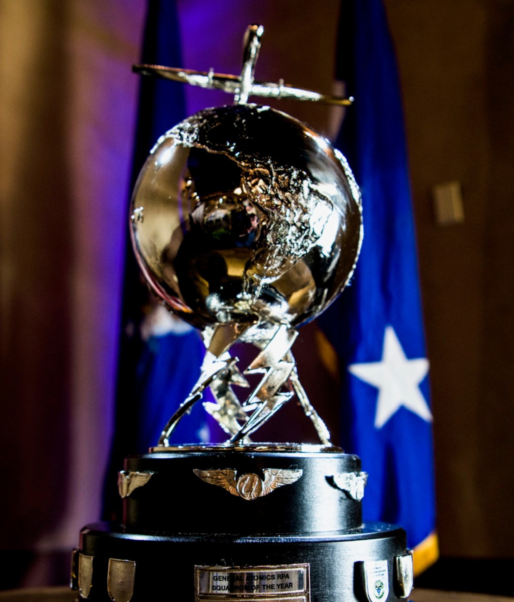 A trophy with a silver globe and lightning bolts displayed on top of 2 black tier circles with military flags draped in the back ground.