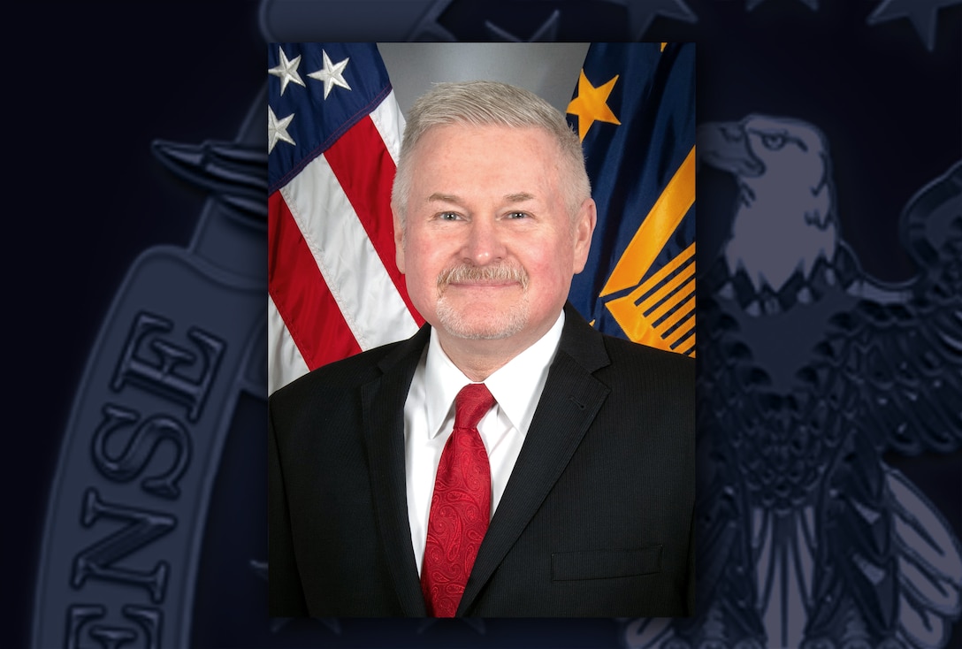 White man in black suit jacket, white shirt and red tie poses in front of the US and DLA flags.