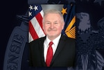 White man in black suit jacket, white shirt and red tie poses in front of the US and DLA flags.