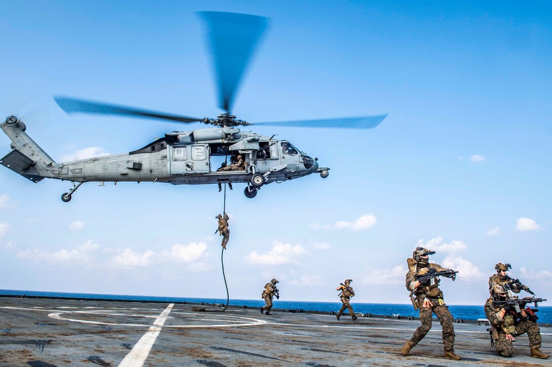 Marines stand on a ship’s deck as other Marines exit a helicopter flying above.