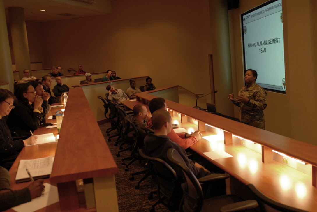 Soldiers in a large room listen to a woman speak.