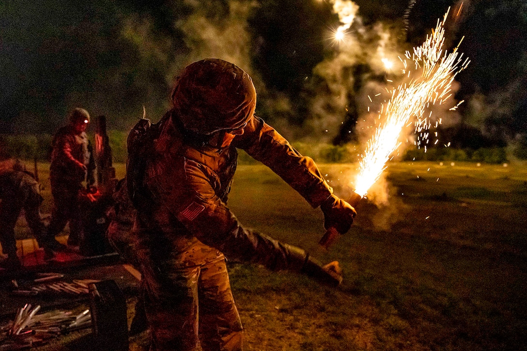 A soldier holds a flaming pyrotechnic flare at a dark range area as others stand nearby.
