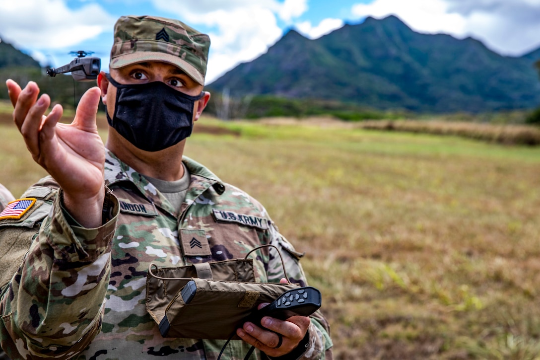 A soldier in a field holds his hand open as a small drone hovers just above it.