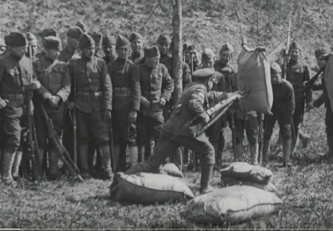 A man stabs a stuffed sack with a bayonet as others watch.