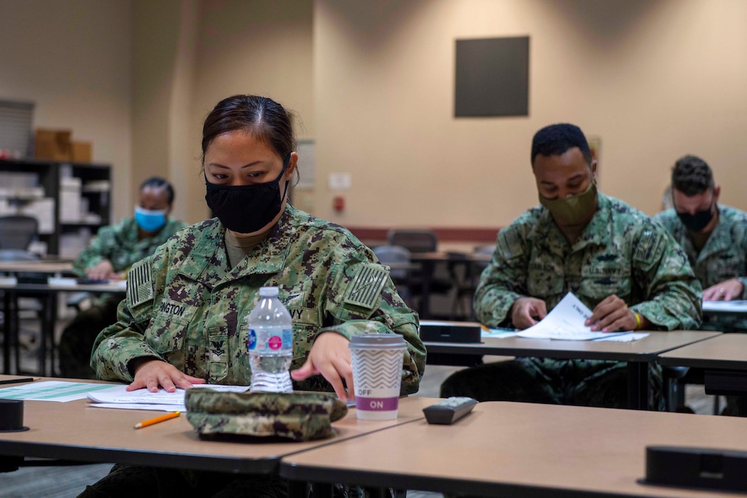 A sailor wearing a face mask sits at a desk; others sit behind.