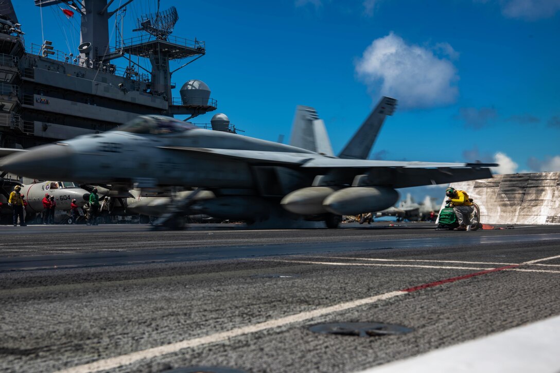 A sailor signals while an aircraft takes off from the deck of a ship.