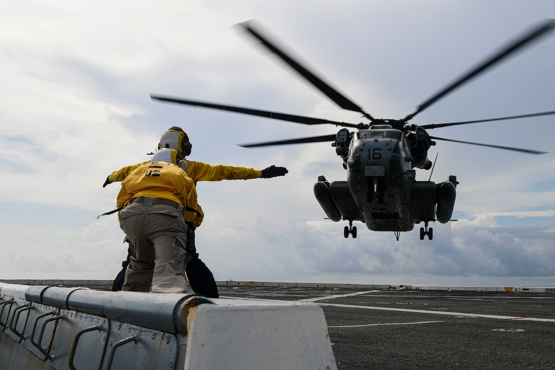 Two sailors in yellow, shown from behind, signal to a helicopter hovering above a flight deck.