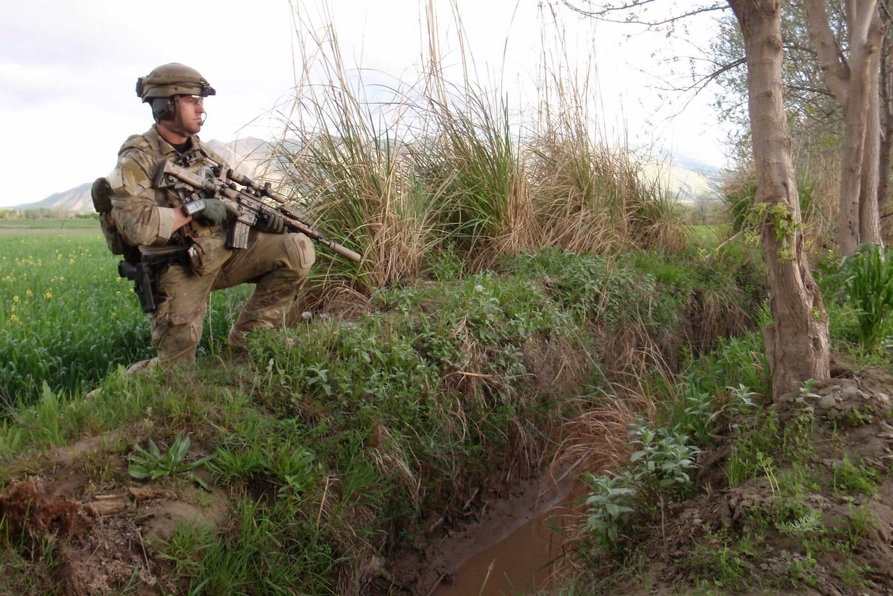 A soldier stands in a field while holding an automatic rifle.