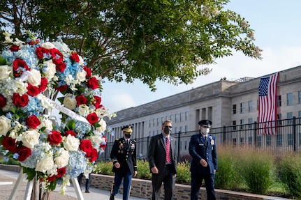 A wreath with red, white and blue flowers stands in the foreground as a large building looms in the background.
