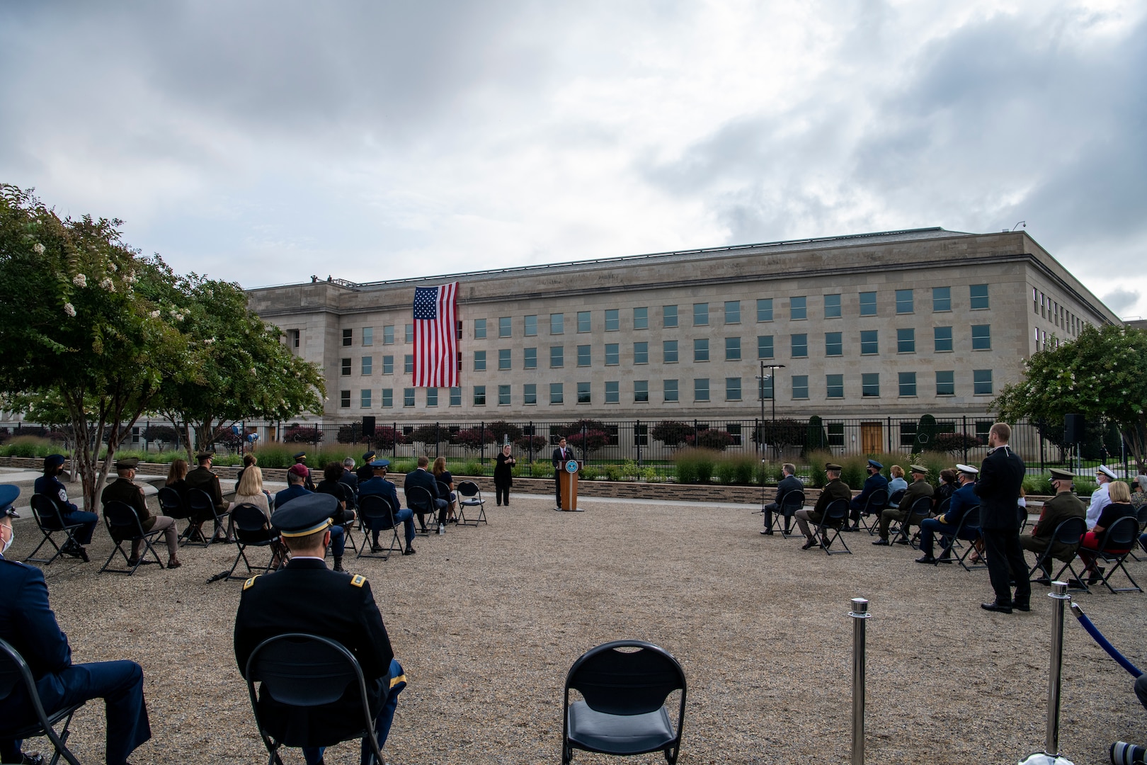 People sit outdoors and listen to someone at a podium; a large building is in the background.