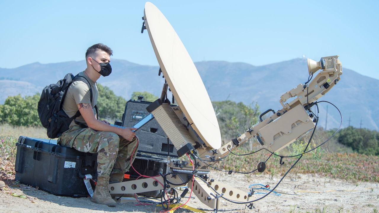 A man in a military uniform sits outside on a large box next to a satellite communications dish.