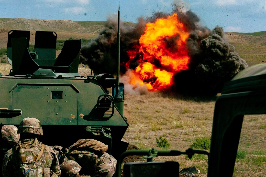 Guardsmen kneel behind a tank with an explosion in the background.