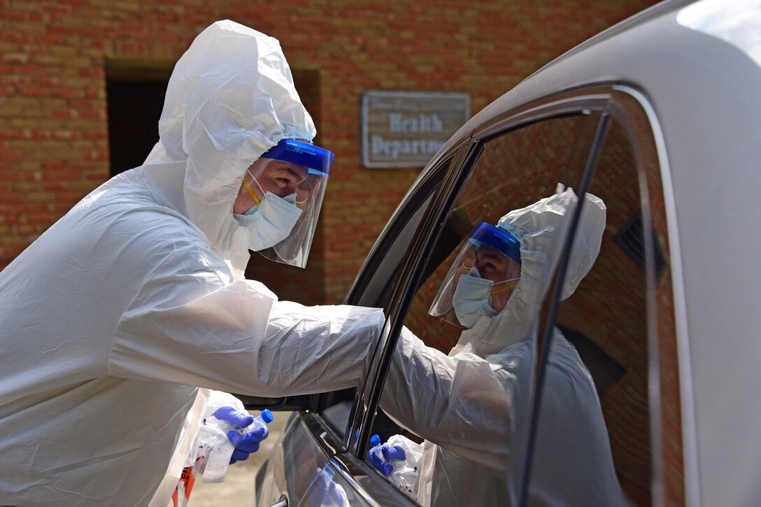 Masked, visored person wearing full protective clothing leans forward and reaches into a vehicle.