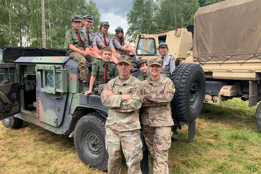 Children in foreign scout uniforms sit atop a military vehicle with their scout master standing nearby. Two soldiers stand in front of the vehicle as all smile for the camera.